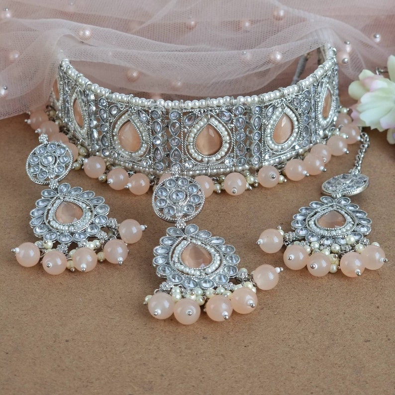 Peut inclure: Un ensemble de bijoux de mari&eacute;e argent&eacute;s comprenant un collier ras du cou, des boucles d'oreilles et un bijou de t&ecirc;te. L'ensemble comprend des pierres en forme de goutte d'eau de couleur p&ecirc;che, de petites perles et des pampilles de perles p&ecirc;che. Les bijoux sont pr&eacute;sent&eacute;s sur un fond neutre.