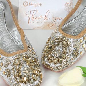 Peut inclure: Une paire de chaussures plates argent&eacute;es orn&eacute;es de strass dor&eacute;s et transparents. Les chaussures ont une bordure en cuir beige et sont d&eacute;cor&eacute;es d'un motif floral. Les chaussures sont sur une surface blanche avec une fleur blanche.