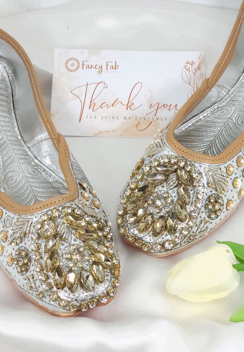 Peut inclure: Une paire de chaussures plates argent&eacute;es orn&eacute;es de strass dor&eacute;s et transparents. Les chaussures ont une bordure en cuir beige et sont d&eacute;cor&eacute;es d'un motif floral. Les chaussures sont sur une surface blanche avec une fleur blanche.