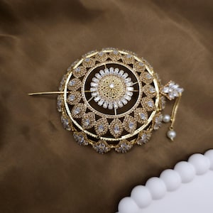 Könnte beinhalten: Ein goldfarbenes Haaraccessoire mit kreisförmigem Design. Die Mitte weist ein detailliertes Filigranmuster auf, das von klaren, oval geformten Steinen umgeben ist. Ein dekorativer Cluster aus Steinen und hängenden Perlen ist angebracht.