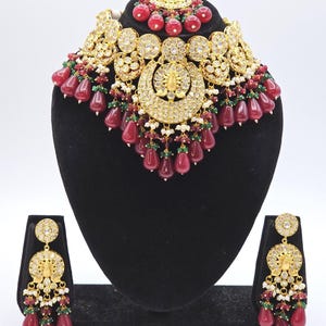 Pu&ograve; includere: Un set di collana e orecchini in oro e bianco con perline rosse e verdi. La collana &egrave; di tipo choker con un grande ciondolo al centro. Gli orecchini sono a goccia con un design simile al ciondolo.