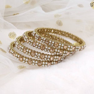 May include: Set of four gold bangles with a delicate design featuring small, clear rhinestones and white pearls.
