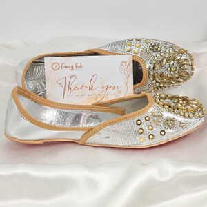 Peut inclure: Une paire de chaussures plates argent&eacute;es orn&eacute;es de d&eacute;tails dor&eacute;s. Les chaussures ont une bordure en cuir beige et sont d&eacute;cor&eacute;es de strass et de perles. Les chaussures sont sur un fond de tissu blanc. L'image comprend &eacute;galement une carte de remerciement avec le texte "Fancy Fab" et "Merci d'&ecirc;tre mon client".