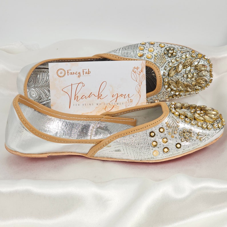 Peut inclure: Une paire de chaussures plates argent&eacute;es orn&eacute;es de d&eacute;tails dor&eacute;s. Les chaussures ont une bordure en cuir beige et sont d&eacute;cor&eacute;es de strass et de perles. Les chaussures sont sur un fond de tissu blanc. L'image comprend &eacute;galement une carte de remerciement avec le texte "Fancy Fab" et "Merci d'&ecirc;tre mon client".