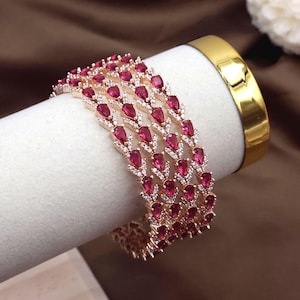 Könnte beinhalten: Vier roségoldene Armreifen mit abwechselnd rubinroten, tropfenförmigen Steinen und kleinen weißen Steinen. Die Armreifen werden auf einer weißen Oberfläche mit einem goldenen Band im Hintergrund präsentiert.
