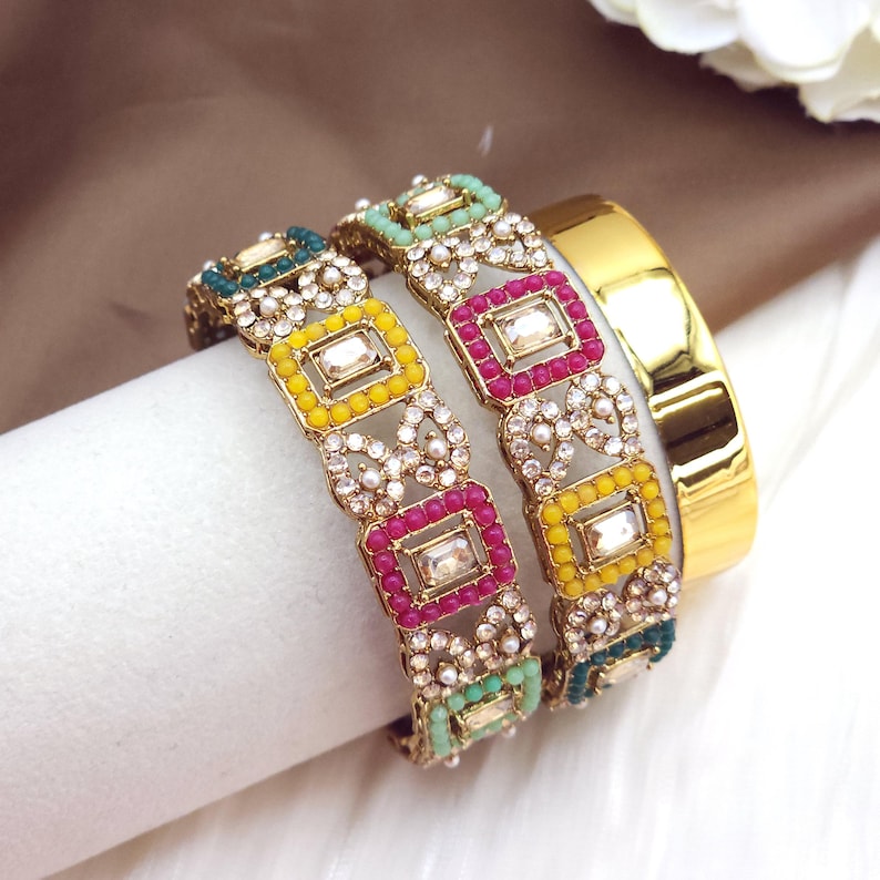 May include: Gold-toned bangles with colorful square accents. The bangles feature yellow, pink, and teal squares, surrounded by small white stones. A plain gold bangle is also visible.