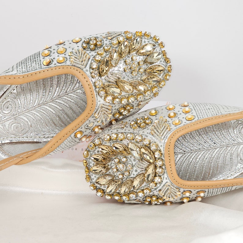 Peut inclure: Une paire de chaussures plates argent&eacute;es orn&eacute;es de strass dor&eacute;s et transparents. Les chaussures ont une pointe pointue et un talon bas.