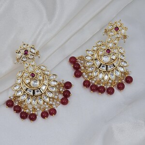 May include: A pair of gold-toned earrings with a floral design. The earrings feature clear and ruby-colored gemstones, along with small white pearls. The bottom of the earrings have a cluster of ruby-colored beads.