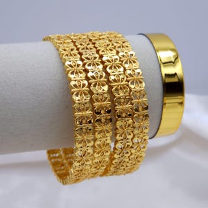 May include: Several gold bangles with detailed floral patterns are displayed. A plain gold band sits beside them. The jewellery is shown against a white backdrop, highlighting the intricate designs and the polished finish of the gold.