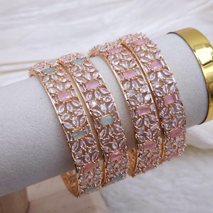 May include: A set of four rose gold-toned bangles featuring a floral pattern of clear marquise-cut stones. Each bangle is accented with rectangular pastel pink and light green stones arranged in a repeating sequence along the metal band.