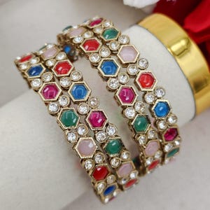 May include: A set of two gold-toned metal bangles featuring a repeating pattern of hexagonal-shaped colourful stones in red, blue, green, and light pink, interspersed with small clear round rhinestones set in a metallic frame.