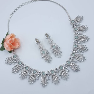 May include: A silver-toned necklace and earring set with a delicate design. The necklace features a series of teardrop-shaped, light-green stones, accented with clear crystals. The matching earrings echo the necklace's design. A peach-colored flower adds a soft contrast.