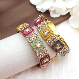 May include: Two gold bangles with colorful square and round bead accents. Each bangle features a central rectangular clear stone surrounded by pink, yellow, green, and blue beads. The bangles are embellished with small clear crystals.