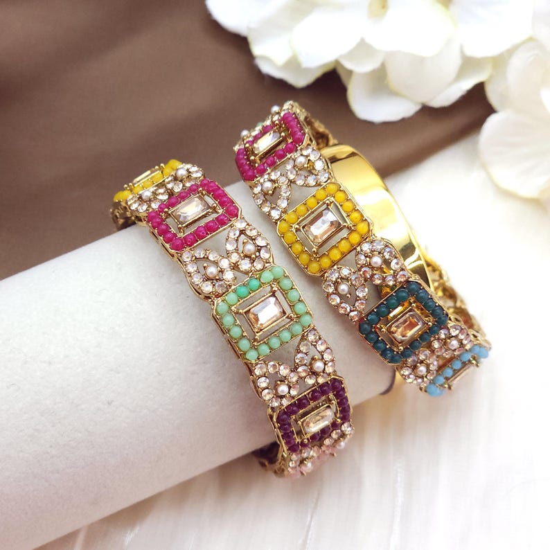 May include: Two gold bangles with colorful square and round bead accents. Each bangle features a central rectangular clear stone surrounded by pink, yellow, green, and blue beads. The bangles are embellished with small clear crystals.