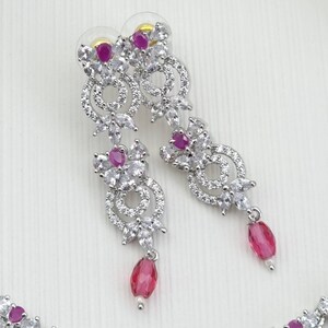 May include: Elegant silver-toned earrings with a floral design. The earrings feature clear, sparkling stones and vibrant pink gemstones. Each earring has a dangling pink teardrop-shaped stone, adding a touch of color and movement to the design.