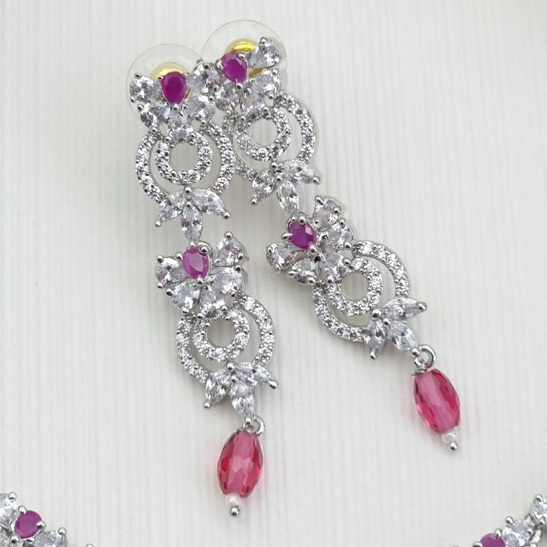 May include: Elegant silver-toned earrings with a floral design. The earrings feature clear, sparkling stones and vibrant pink gemstones. Each earring has a dangling pink teardrop-shaped stone, adding a touch of color and movement to the design.