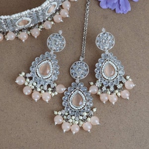 Peut inclure: Un ensemble de bijoux argent&eacute;s comprenant un collier ras du cou, des boucles d'oreilles et un bijou de t&ecirc;te. L'ensemble comprend des pierres en forme de goutte de couleur p&ecirc;che, de petites perles blanches et des pampilles de perles p&ecirc;che. Les bijoux sont con&ccedil;us avec des d&eacute;tails complexes et une esth&eacute;tique vintage.