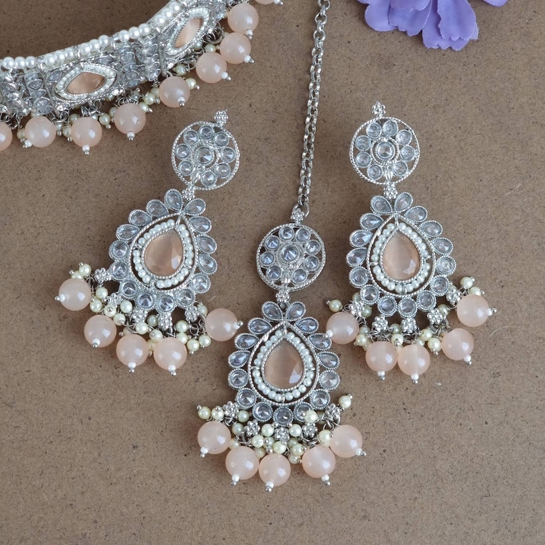Peut inclure: Un ensemble de bijoux argent&eacute;s comprenant un collier ras du cou, des boucles d'oreilles et un bijou de t&ecirc;te. L'ensemble comprend des pierres en forme de goutte de couleur p&ecirc;che, de petites perles blanches et des pampilles de perles p&ecirc;che. Les bijoux sont con&ccedil;us avec des d&eacute;tails complexes et une esth&eacute;tique vintage.