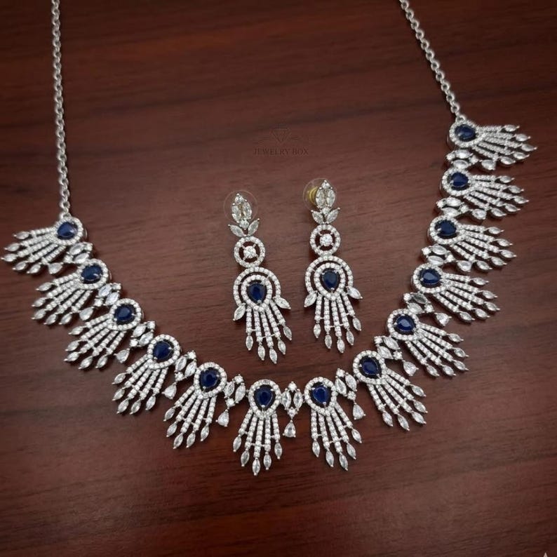 May include: A silver-toned jewelry set featuring a necklace and matching earrings. The necklace and earrings have a repeating design of white crystals and dark blue gemstones. The jewelry is displayed on a dark brown wooden surface.