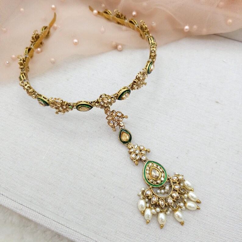 May include: Gold-toned headpiece with floral and teardrop designs. The headpiece features green enamel accents and is adorned with clear crystals and faux pearls. The design includes a central pendant with a teardrop shape and dangling pearls.