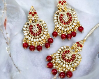 May include: A set of gold-tone earrings and a matching tikka, all adorned with red and white stones. The earrings and tikka feature a circular design with a central red stone surrounded by smaller white stones. The earrings have a dangling chain with red beads.
