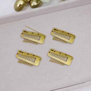 Puede incluir: Cuatro pinzas para el pelo de color dorado con un diseño de peine y un pequeño pasador en cada pinza. Las pinzas están dispuestas en dos filas de dos.