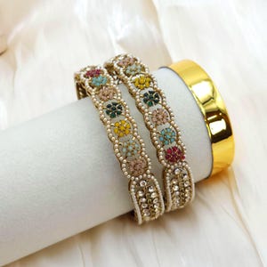 May include: Two gold-toned bangles with colorful floral designs and faux pearls. The bangles are adorned with red, pink, yellow, green, and blue accents. A solid gold bangle is also visible.