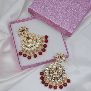 May include: Gold-toned chandelier earrings with clear and red gemstones, and small white pearls. The earrings are displayed in a pink glitter box on a white satin surface. The earrings have a floral design.
