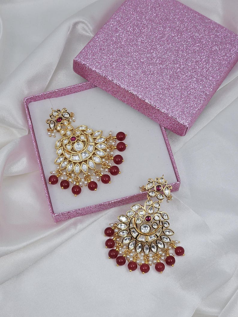 May include: Gold-toned chandelier earrings with clear and red gemstones, and small white pearls. The earrings are displayed in a pink glitter box on a white satin surface. The earrings have a floral design.