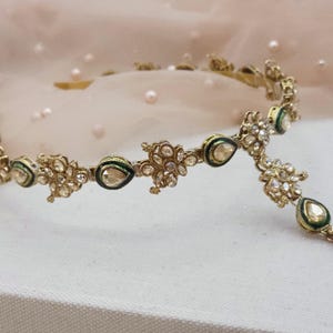 May include: A gold-toned headpiece with intricate floral and teardrop designs. The headpiece features clear gemstones and green and black enamel accents. The delicate jewelry is likely for a special occasion.