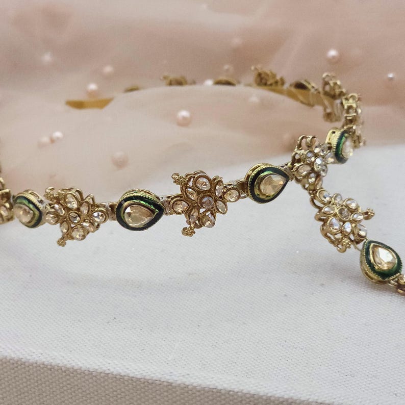 May include: A gold-toned headpiece with intricate floral and teardrop designs. The headpiece features clear gemstones and green and black enamel accents. The delicate jewelry is likely for a special occasion.