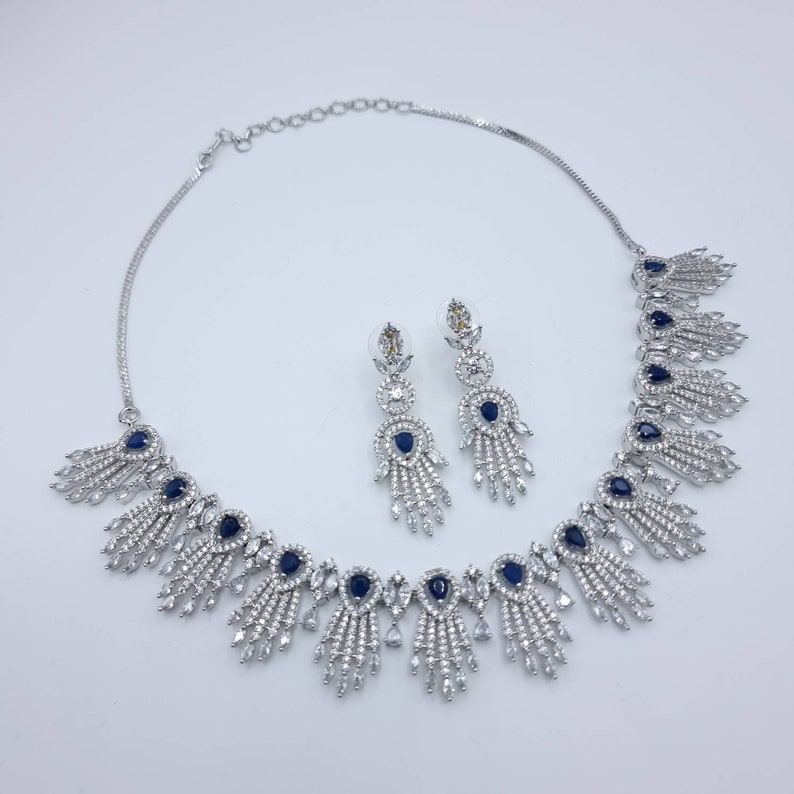 May include: A silver-toned jewelry set featuring a necklace and matching earrings. The necklace and earrings have a repeating design of clear and dark blue stones. The necklace is approximately 16 inches long.