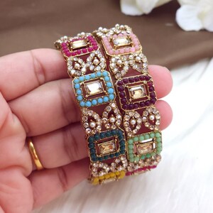 May include: Two ornate bangles with a gold-tone metal base. Each bangle features square, colorful gemstone settings in pink, blue, green, and purple, accented with small white pearls and clear crystals. The bangles are designed with a vintage aesthetic.