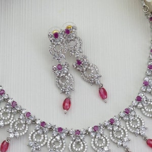 May include: A silver-toned jewelry set featuring a necklace and matching earrings. The set is adorned with clear, sparkling stones and vibrant pink gemstones. The earrings have a dangling teardrop design, and the necklace has a similar pendant style.