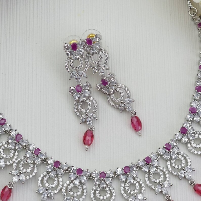 May include: A silver-toned jewelry set featuring a necklace and matching earrings. The set is adorned with clear, sparkling stones and vibrant pink gemstones. The earrings have a dangling teardrop design, and the necklace has a similar pendant style.