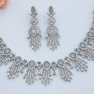 May include: A silver-toned jewelry set featuring a necklace and matching earrings. The necklace and earrings have a delicate design with clear and pale green gemstones. The earrings are long and dangle, while the necklace has a symmetrical pattern.