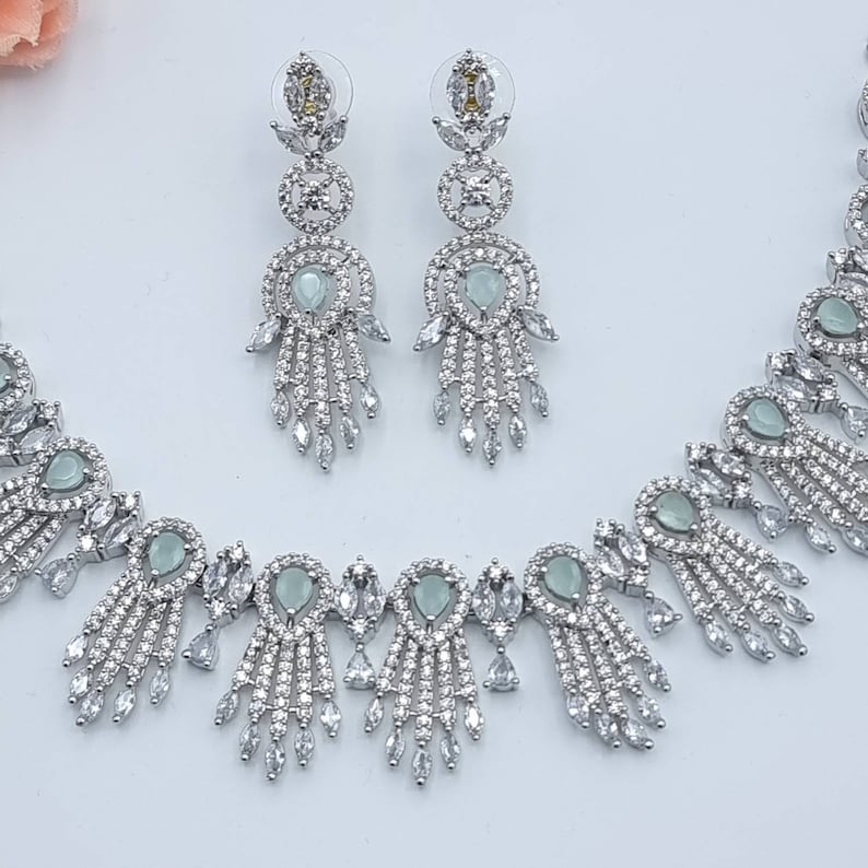 May include: A silver-toned jewelry set featuring a necklace and matching earrings. The necklace and earrings have a delicate design with clear and pale green gemstones. The earrings are long and dangle, while the necklace has a symmetrical pattern.