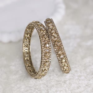 May include: Two gold-colored bangles with intricate detailing. The bangles are adorned with small, sparkling gemstones and geometric patterns. One bangle is upright, while the other rests at an angle, showcasing the design.