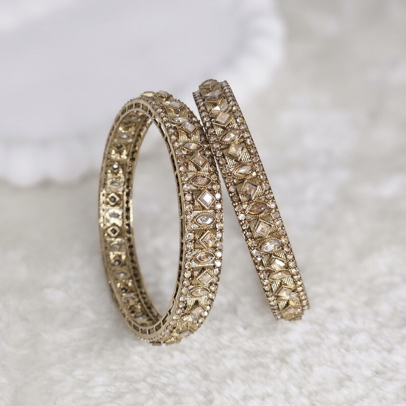 May include: Two gold-colored bangles with intricate detailing. The bangles are adorned with small, sparkling gemstones and geometric patterns. One bangle is upright, while the other rests at an angle, showcasing the design.