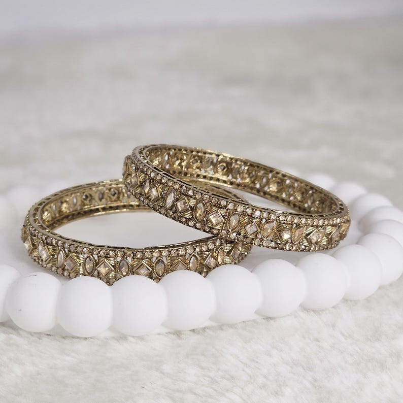 May include: Two gold-toned bangles with intricate detailing and embedded gemstones. The bangles are displayed on a white, beaded surface, creating a contrast between the warm metal and the cool white backdrop. The jewelry is designed with geometric and floral patterns.