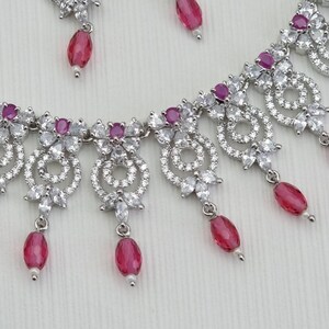 May include: A silver-toned necklace and earring set with clear and ruby-colored gemstones. The necklace features a floral and circular design with dangling ruby-colored teardrop-shaped pendants. The earrings have a similar design with a single ruby-colored pendant.