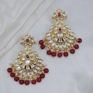 May include: A pair of gold-toned chandelier earrings. Each earring features a central ruby-colored stone, surrounded by clear, faceted stones and small white pearls. The earrings have a crescent-shaped design with ruby-colored bead dangles.