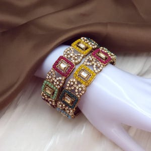 May include: Two ornate gold bangles with colorful square and rectangular gemstones. The bangles feature red, yellow, green, and blue accents, with a delicate, openwork design. The gemstones are set in a gold frame.