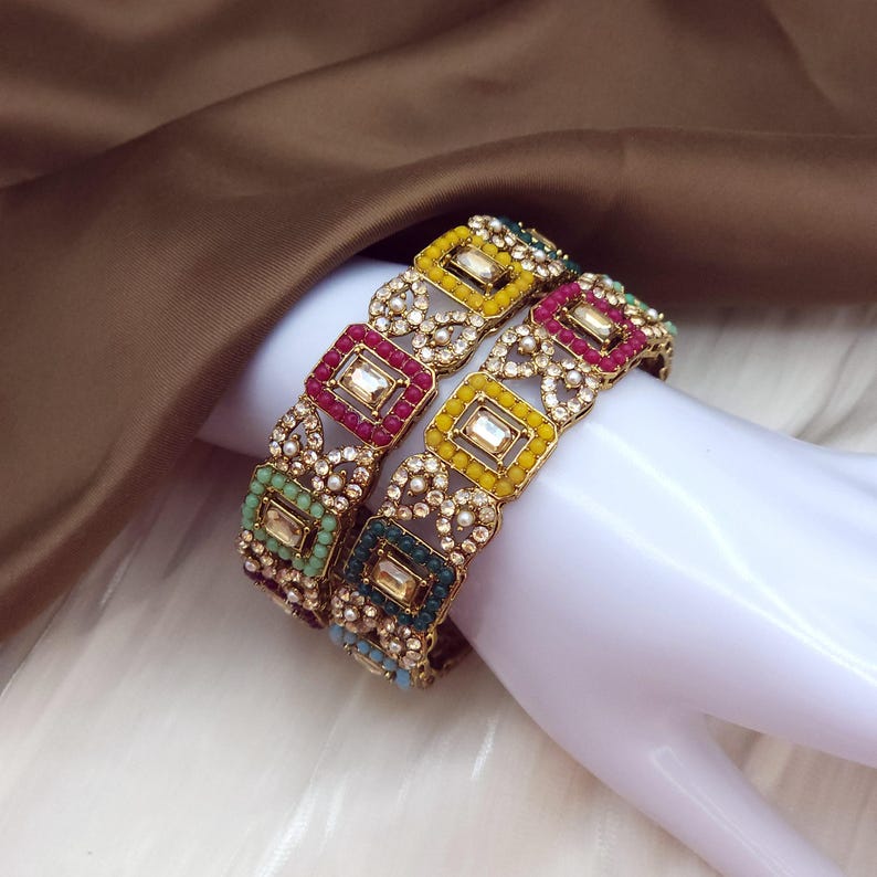 May include: Two ornate gold bangles with colorful square and rectangular gemstones. The bangles feature red, yellow, green, and blue accents, with a delicate, openwork design. The gemstones are set in a gold frame.