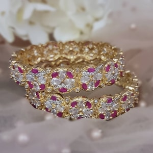 May include: Two gold bangles featuring white and pink stones. The bangles are adorned with a floral design and are stacked on top of each other.