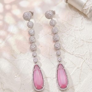 May include: A pair of long drop earrings. Each earring has a series of small, sparkling, round embellishments leading to a teardrop-shaped pink gemstone. The earrings are set in a silver-toned metal. Displayed on a white, textured surface.