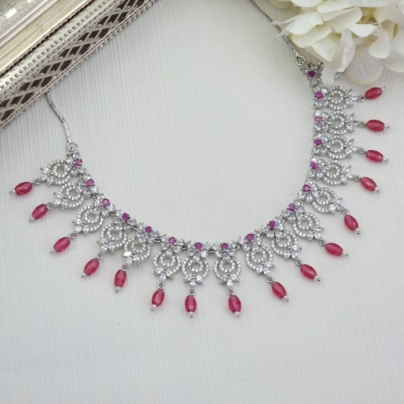 May include: A silver-toned necklace with a floral design, adorned with clear crystals and oval-shaped, ruby-red gemstones. The necklace features a delicate chain and is displayed on a white surface.