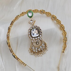 May include: Gold-toned headband with a central pendant featuring a large, square, clear gemstone surrounded by smaller clear stones. The pendant is connected to a smaller green and white gemstone. Small white pearls dangle from the bottom.