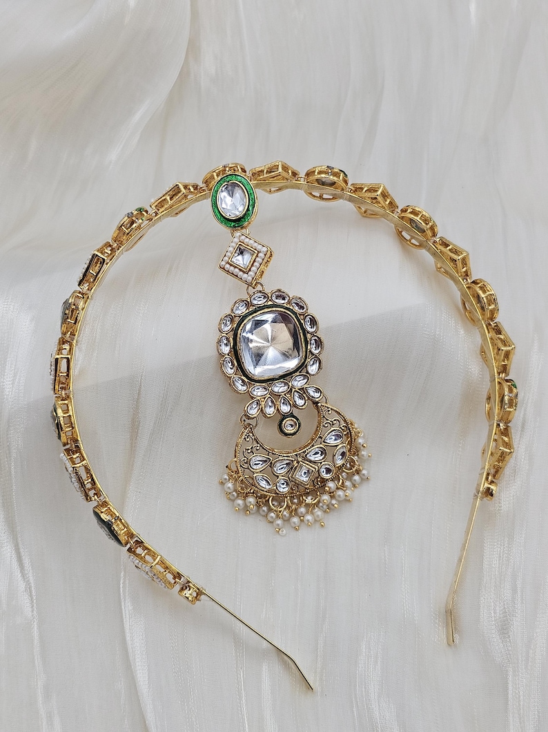 May include: Gold-toned headband with a central pendant featuring a large, square, clear gemstone surrounded by smaller clear stones. The pendant is connected to a smaller green and white gemstone. Small white pearls dangle from the bottom.