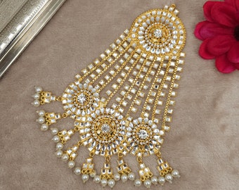May include: A gold and white ornate headpiece with multiple tiers of dangling chains and pearl accents. The headpiece features a large circular design with a central rhinestone.